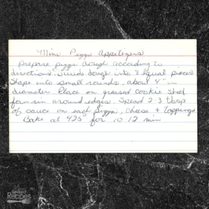 A handwritten recipe card with blue lines and cursive writing on a white index card. It sits on a black marble countertop. The card provides a recipe for mini pizza appetizers, including instructions on preparing dough, shaping rounds, adding sauce, cheese, toppings, and baking.