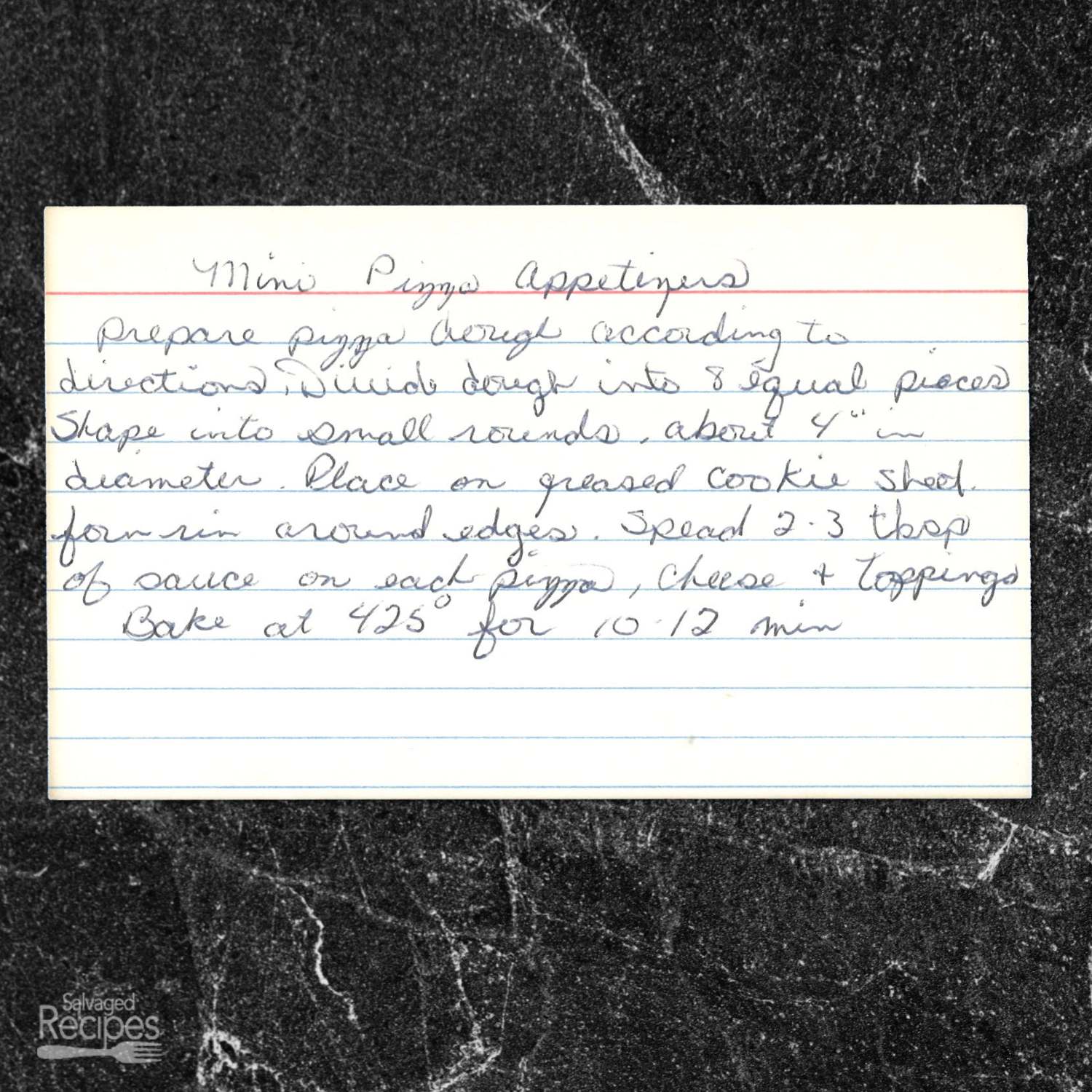A handwritten recipe card with blue lines and cursive writing on a white index card. It sits on a black marble countertop. The card provides a recipe for mini pizza appetizers, including instructions on preparing dough, shaping rounds, adding sauce, cheese, toppings, and baking.