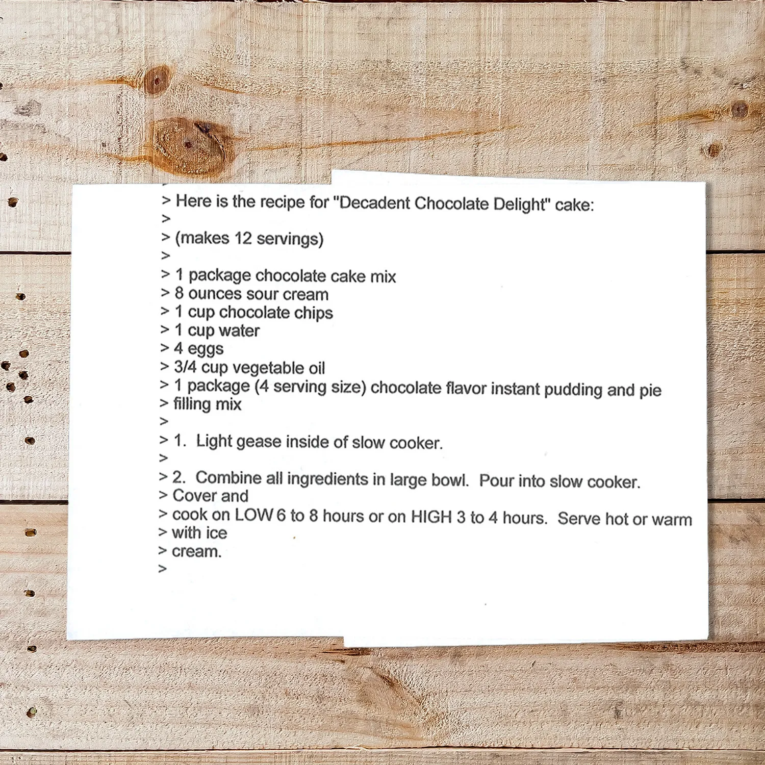 A printed recipe card on a wooden surface featuring the "Decadent Chocolate Delight" cake recipe. The text lists ingredients like chocolate cake mix, sour cream, chocolate chips, and eggs, followed by step-by-step instructions for making the cake in a slow cooker.