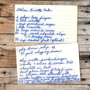 Two vintage, handwritten recipe cards on lined paper are placed on a rustic wooden surface. The top card lists ingredients for "Italian Ricotta Cake" in blue ink, and the second card contains preparation instructions and additional ingredients in matching handwriting.